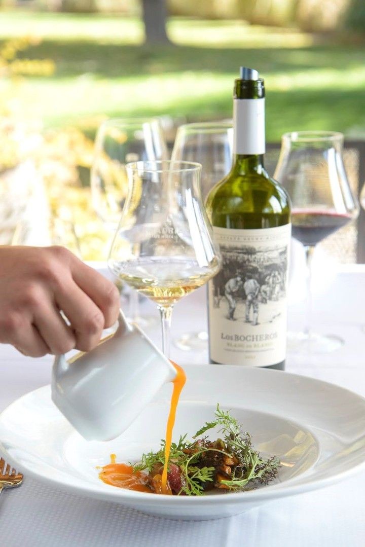 Person pouring orange sauce over a gourmet dish on a table with wine glasses and a bottle.