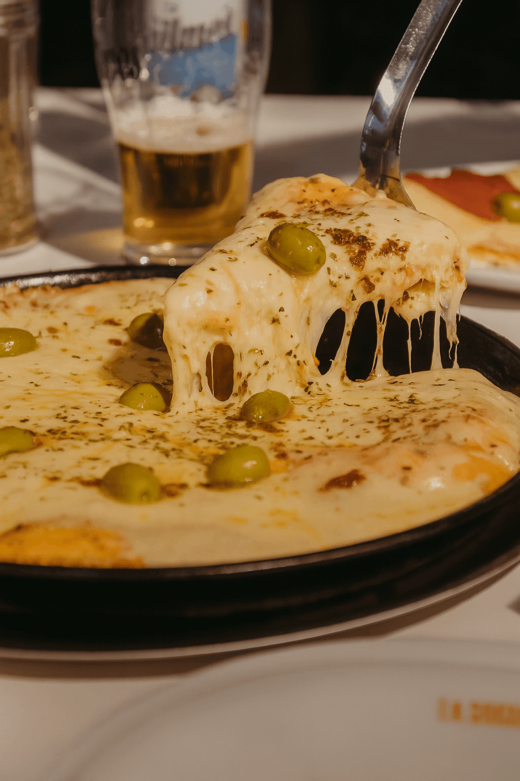Close-up of cheesy pizza slice with olives being lifted, beer glass in background.