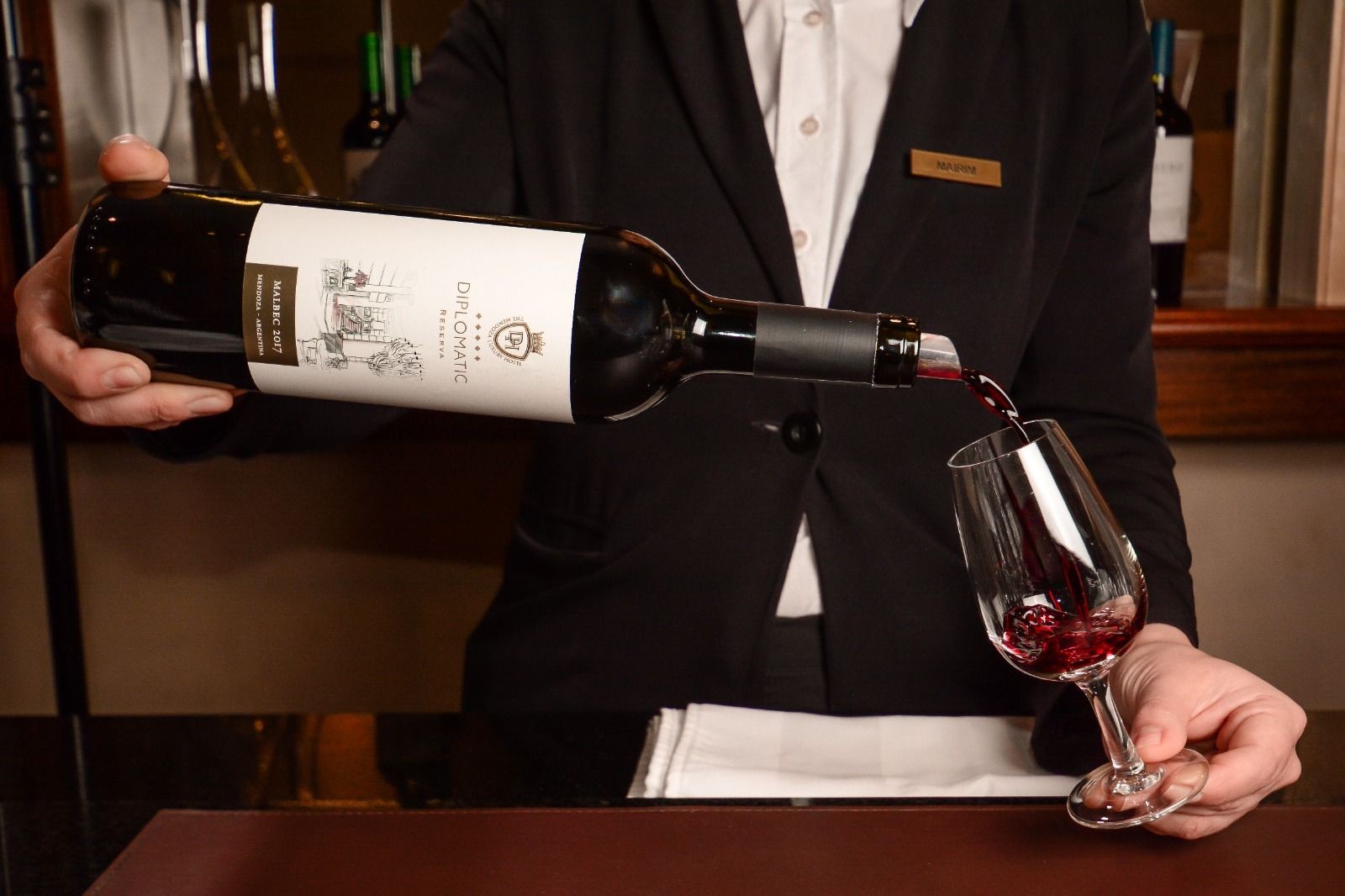 Person in a suit pouring red wine from a bottle labeled 'Diplomatic' into a glass.