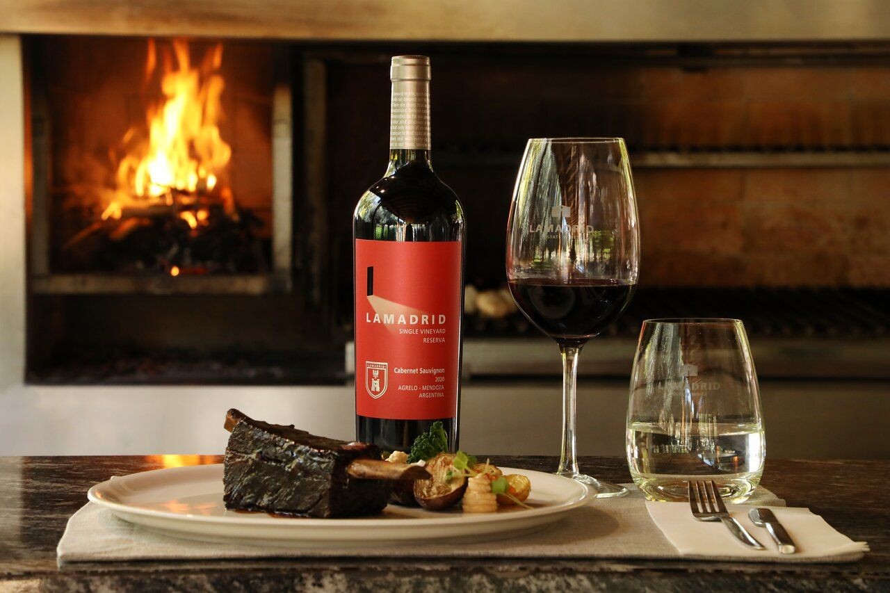 Bottle of Lamadrid wine with a glass, plate of food, and fireplace in the background.